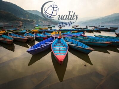 Barche di legno colorate disposte in cerchio su un tranquillo lago nepalese con montagne sullo sfondo, perfette per un tour panoramico. Logo sovrapposto in alto.