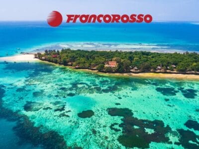 Vista aerea di un'isola tropicale con acqua turchese, barriere coralline e il logo "Francorosso" in alto, che cattura la bellezza di Mare e il fascino di Tanzania e Mare.