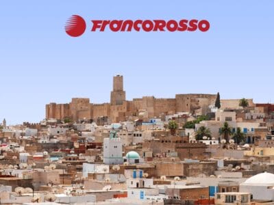 Paesaggio urbano con fortezza storica e numerosi edifici bianchi in Tunisia, il logo Francorosso in alto contro un cielo limpido: scopri Autentica Tunisia.