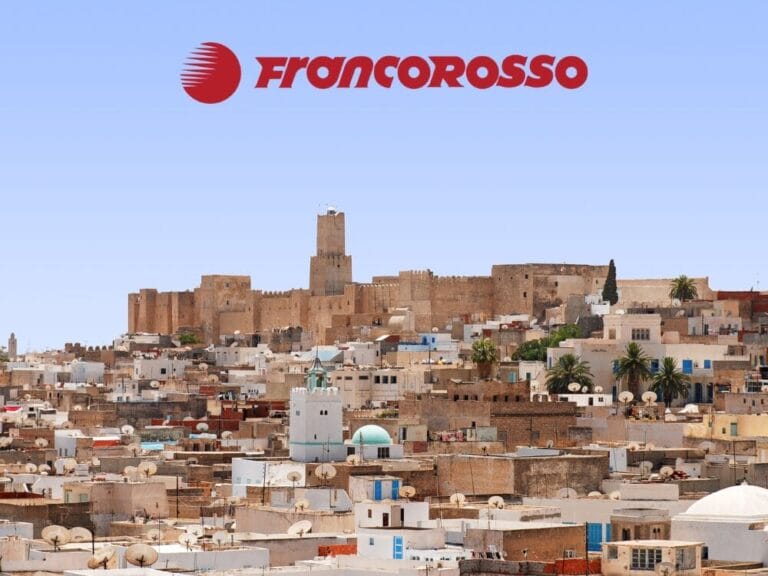 Paesaggio urbano con fortezza storica e numerosi edifici bianchi in Tunisia, il logo Francorosso in alto contro un cielo limpido: scopri Autentica Tunisia.