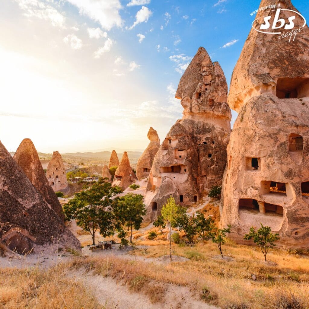 Formazioni rocciose a forma di cono con abitazioni scolpite al tramonto in Cappadocia, Turchia, con alcuni alberi verdi.