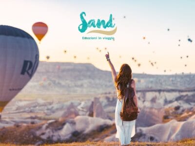 Una donna con uno zaino indica le mongolfiere che sorvolano un paesaggio roccioso all'alba.