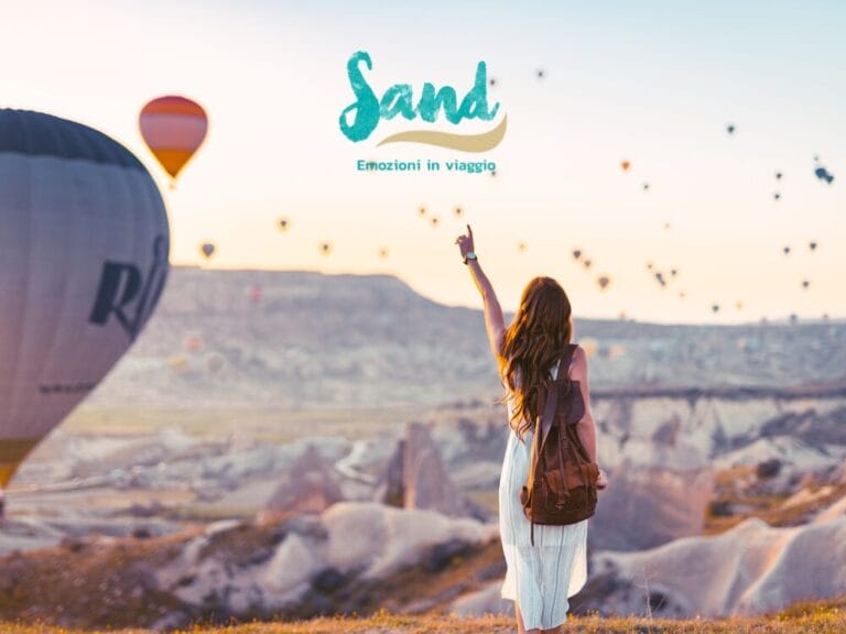 Una donna con uno zaino indica le mongolfiere che sorvolano un paesaggio roccioso all'alba.