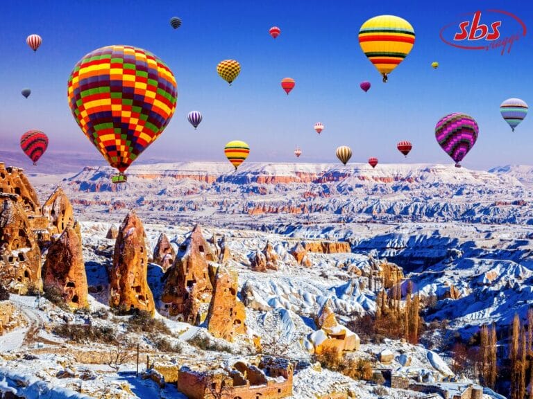 Coloratissime mongolfiere fluttuano sul paesaggio roccioso e innevato della Cappadocia sotto un cielo azzurro e limpido: una scena magica in qualsiasi tour della Turchia o in un indimenticabile viaggio invernale.