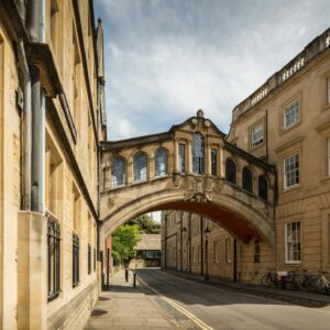 Un ponte di pietra con finestre ad arco collega due edifici storici su una strada tranquilla di Oxford, in Inghilterra, una scena stimolante per chi desidera studiare nel Regno Unito. Diverse biciclette sono parcheggiate nelle vicinanze sotto un cielo parzialmente nuvoloso.
