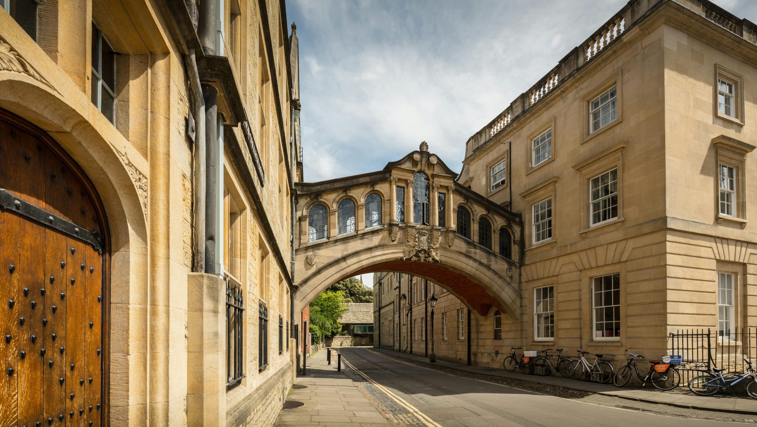 Un ponte di pietra con finestre ad arco collega due edifici storici su una strada tranquilla di Oxford, in Inghilterra, una scena stimolante per chi desidera studiare nel Regno Unito. Diverse biciclette sono parcheggiate nelle vicinanze sotto un cielo parzialmente nuvoloso.