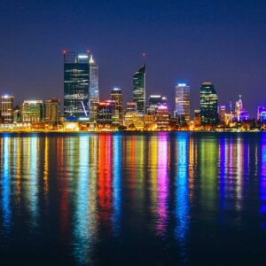 Vista notturna dello skyline di Perth in Australia, con alti edifici illuminati da luci colorate che si riflettono vivacemente sull'acqua calma. Il cielo limpido e blu scuro esalta l'atmosfera luminosa della città, perfetta per chi vuole studiare Perth.