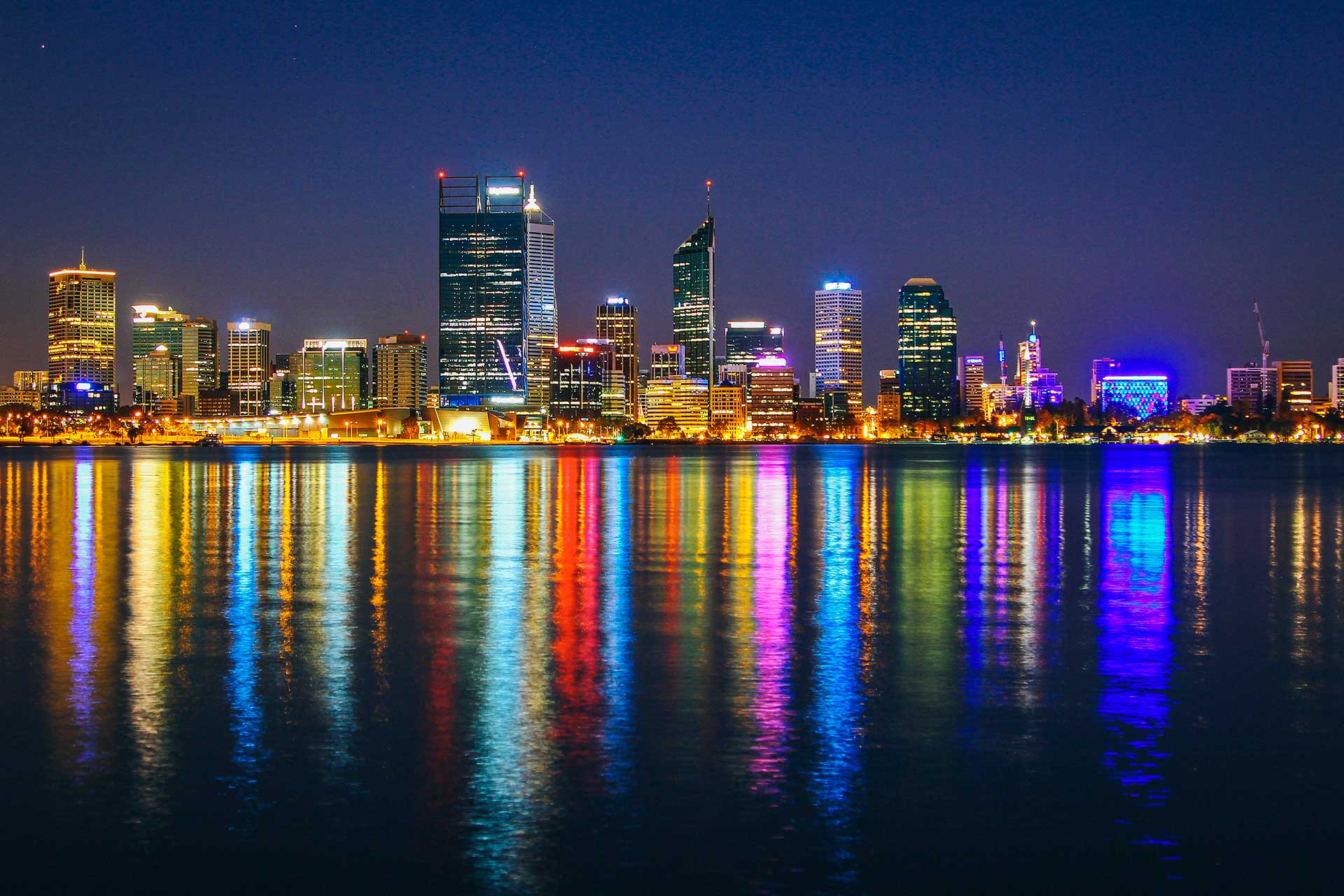 Vista notturna dello skyline di Perth in Australia, con alti edifici illuminati da luci colorate che si riflettono vivacemente sull'acqua calma. Il cielo limpido e blu scuro esalta l'atmosfera luminosa della città, perfetta per chi vuole studiare Perth.