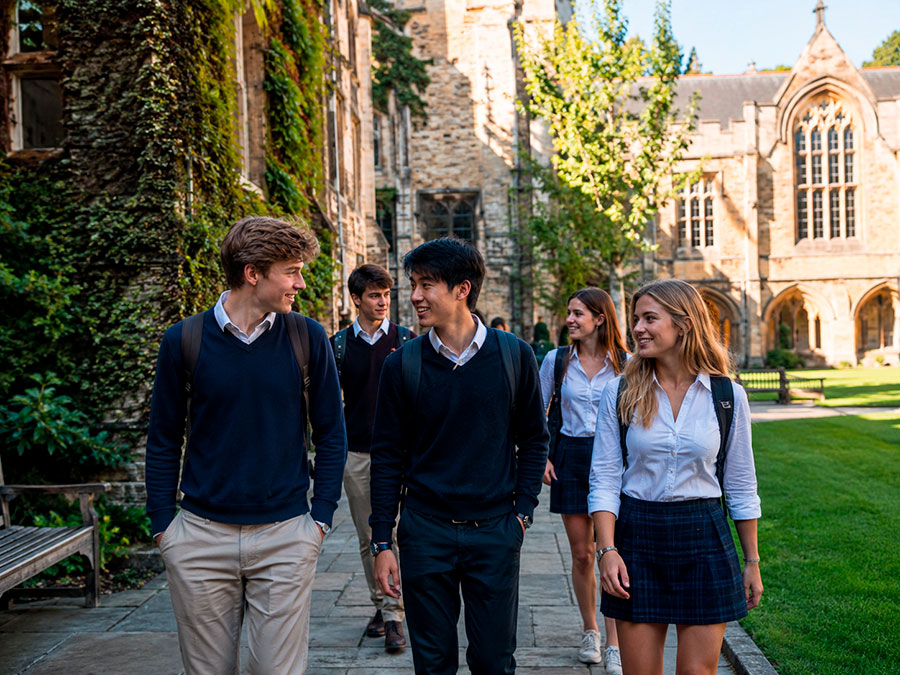 Un gruppo di studenti in uniforme scolastica cammina insieme in una giornata di sole attraverso un campus verdeggiante con edifici in pietra, sorridendo e parlando.