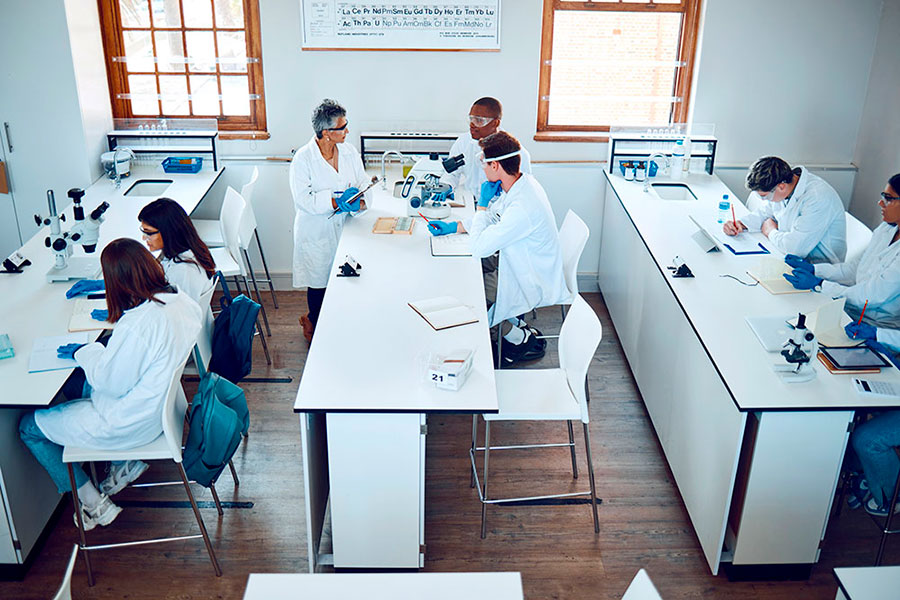 Un gruppo di scienziati in camice bianco lavora su tavoli da laboratorio con microscopi, quaderni e campioni, impegnandosi in esperimenti e discussioni in un'aula luminosa e ben illuminata.