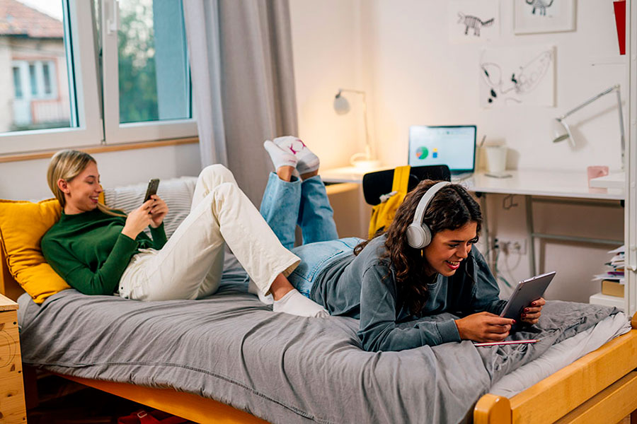 Due giovani donne si rilassano in un'accogliente camera da letto; una è sdraiata sul letto e utilizza uno smartphone, mentre l'altra, con le cuffie, sorride e utilizza un tablet. Sullo sfondo una scrivania con un computer.