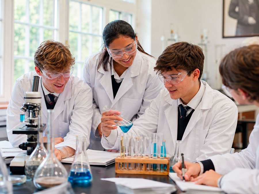 Quattro studenti in camice e occhiali di sicurezza lavorano insieme al tavolo di un laboratorio di scienze, esaminando provette con liquido blu, usando quaderni e sorridendo, con un microscopio e becher nelle vicinanze.