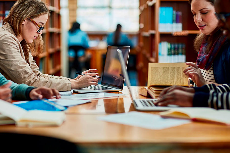 Diverse persone siedono al tavolo di una biblioteca per studiare, utilizzando computer portatili e libri. Sullo sfondo ci sono scaffali di libri e l'ambiente appare concentrato e tranquillo.