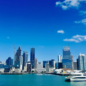 Skyline di una città moderna con alti edifici sotto un cielo blu, vista dall'acqua. Una barca bianca è attraccata nel porto turchese in primo piano.