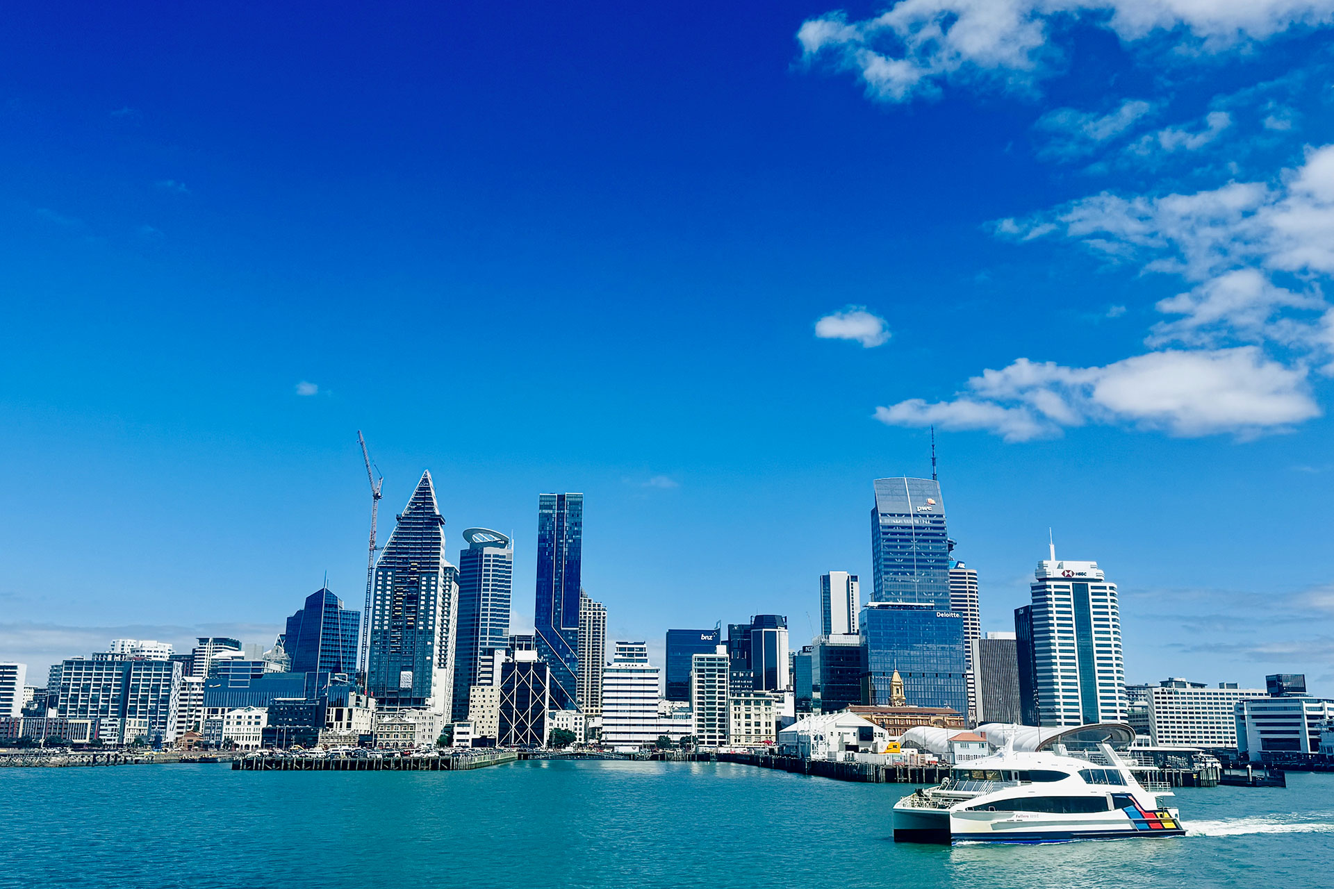 Skyline di una città moderna con alti edifici sotto un cielo blu, vista dall'acqua. Una barca bianca è attraccata nel porto turchese in primo piano.