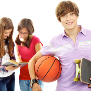 Un ragazzo adolescente con in mano un pallone da basket e uno skateboard sorride alla telecamera, mentre due ragazze sullo sfondo leggono un libro insieme, con un'aria rilassata e disinvolta.