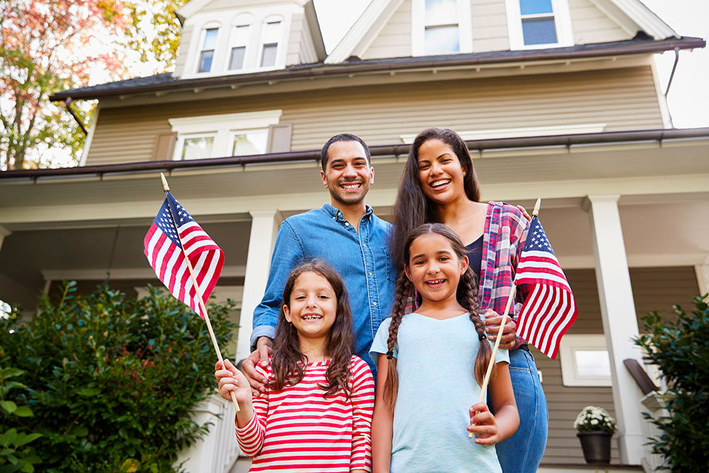 Una famiglia sorridente di quattro persone si trova all'esterno di una casa beige con rifiniture bianche, circondata dal verde. I bambini tengono in mano le bandiere americane, catturando una classica scena da liceo americano. È giorno e la famiglia sembra felice.