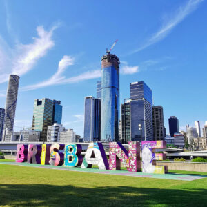 Grandi lettere colorate con la scritta "BRISBANE" campeggiano sull'erba, con grattacieli della città moderna e un edificio in costruzione sullo sfondo, sotto un cielo blu brillante con nuvole a batuffolo.