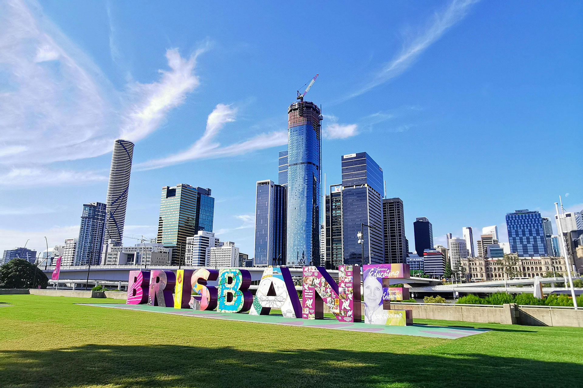 Grandi lettere colorate con la scritta "BRISBANE" campeggiano sull'erba, con grattacieli della città moderna e un edificio in costruzione sullo sfondo, sotto un cielo blu brillante con nuvole a batuffolo.