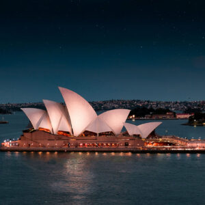 L'Opera House di Sydney è illuminata di notte, con i suoi iconici tetti bianchi a forma di vela che si riflettono nel porto e le luci della città visibili sullo sfondo sotto un cielo stellato.