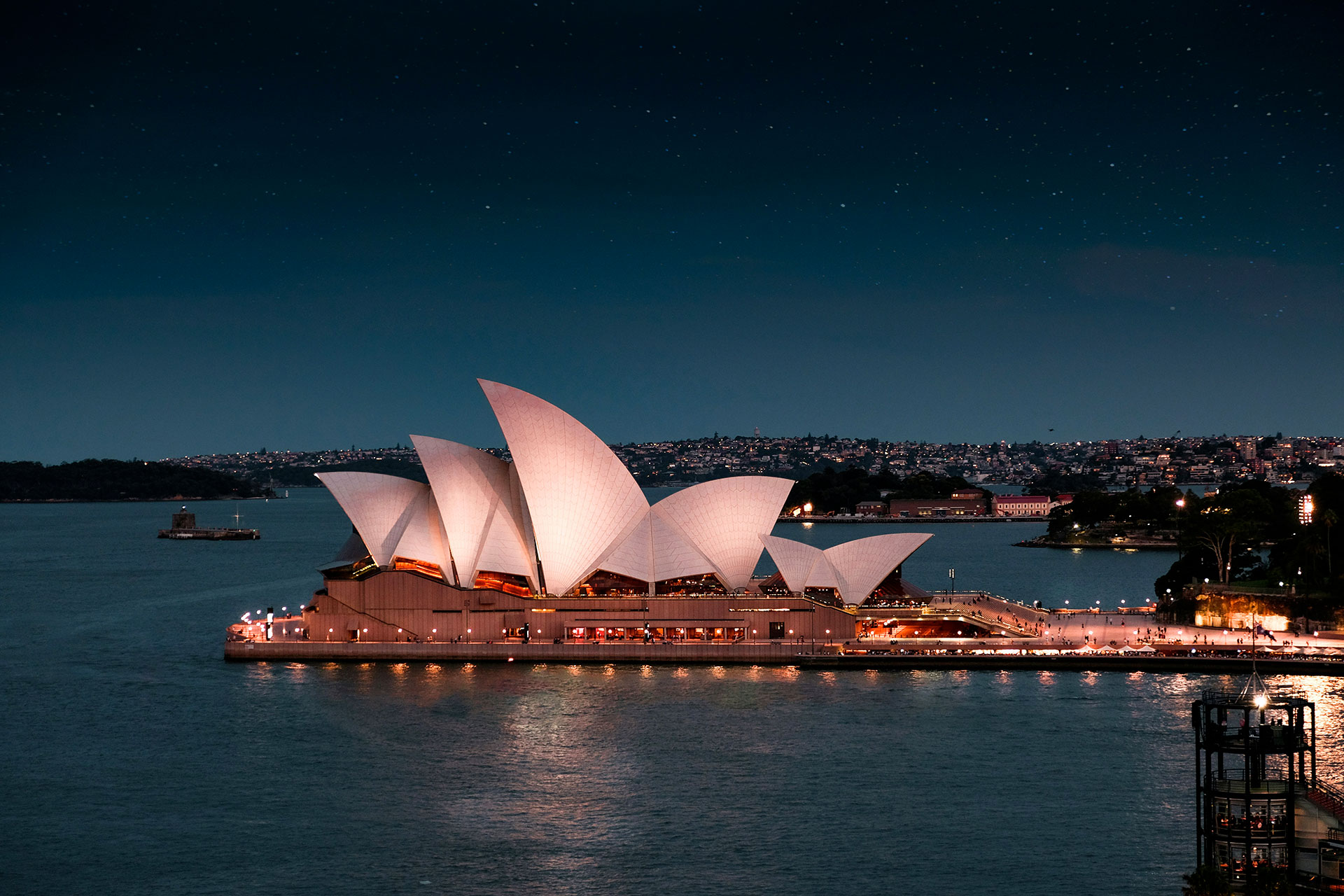 L'Opera House di Sydney è illuminata di notte, con i suoi iconici tetti bianchi a forma di vela che si riflettono nel porto e le luci della città visibili sullo sfondo sotto un cielo stellato.