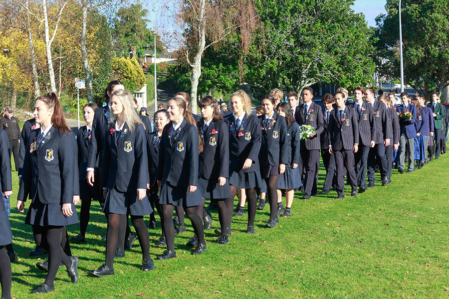 Un gruppo di studenti in giacca e cravatta scura e gonna o pantaloni cammina in file ordinate attraverso un campo erboso, alcuni portando fiori: uno spaccato della vita studentesca di chi sceglie di studiare un anno in Nuova Zelanda.