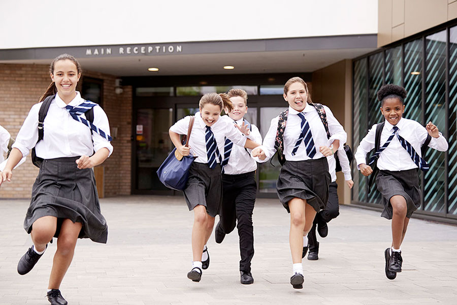 Un gruppo di scolari sorridenti in uniforme corre eccitato fuori da un edificio scolastico, ansioso di iniziare l'anno scolastico in Nuova Zelanda, con un cartello che recita "Main Reception" sopra l'ingresso.