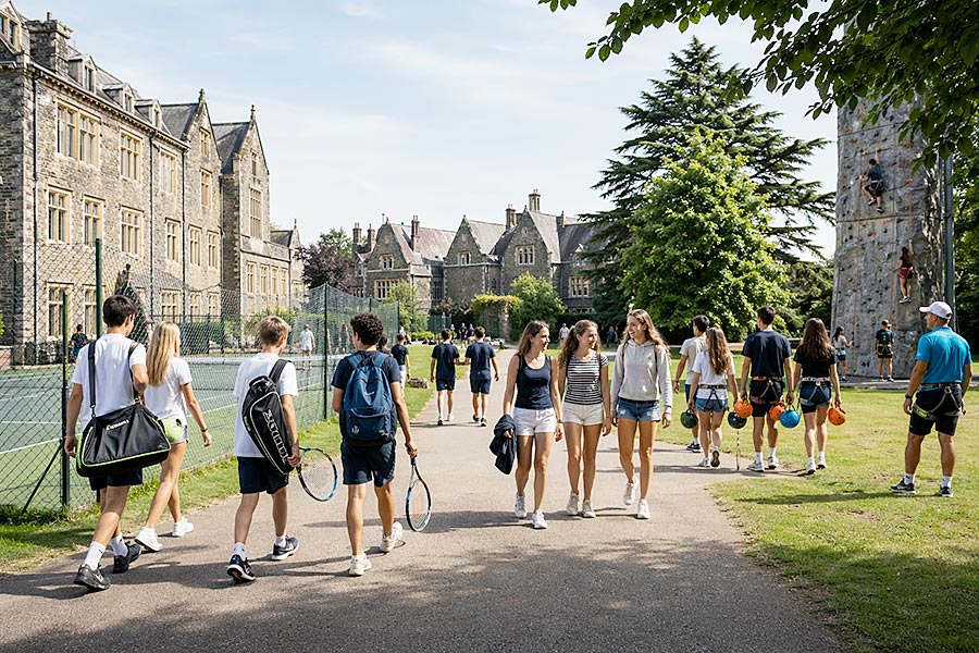 Studenti in tenuta sportiva camminano lungo un sentiero accanto a campi da tennis ed edifici scolastici storici, mentre altri trasportano l'attrezzatura da arrampicata verso una parete da arrampicata. La scena vivace cattura l'eccitazione di un campo multiattività in una giornata di sole.