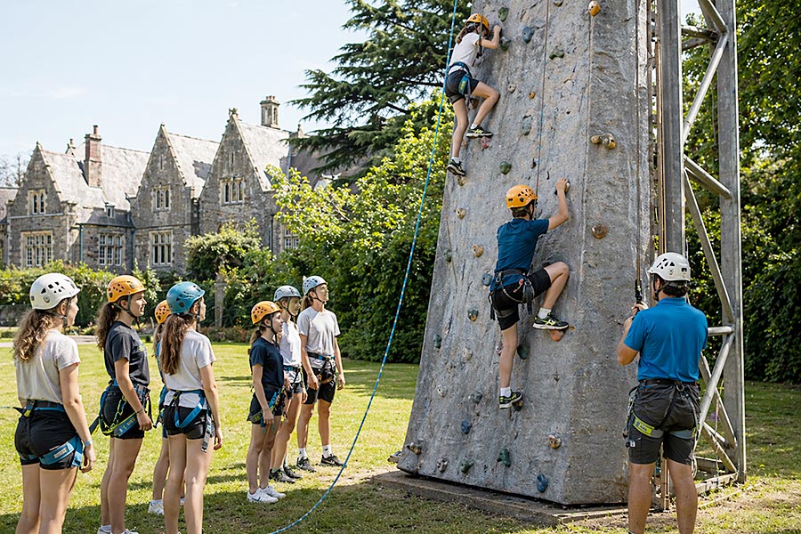Un gruppo di giovani di un campo avventura, con caschi e imbracature, osserva due scalatori che salgono su una parete di arrampicata all'aperto sotto la supervisione di un istruttore, con un grande edificio in pietra sullo sfondo.