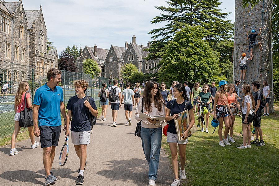 Studenti e adulti camminano e parlano all'esterno di un grande edificio scolastico in pietra in una giornata di sole al Campo Multiattività. Alcuni tengono in mano attrezzature sportive e materiale artistico, mentre altri si arrampicano su una parete di roccia lì vicino. La scena è vivace e sociale.