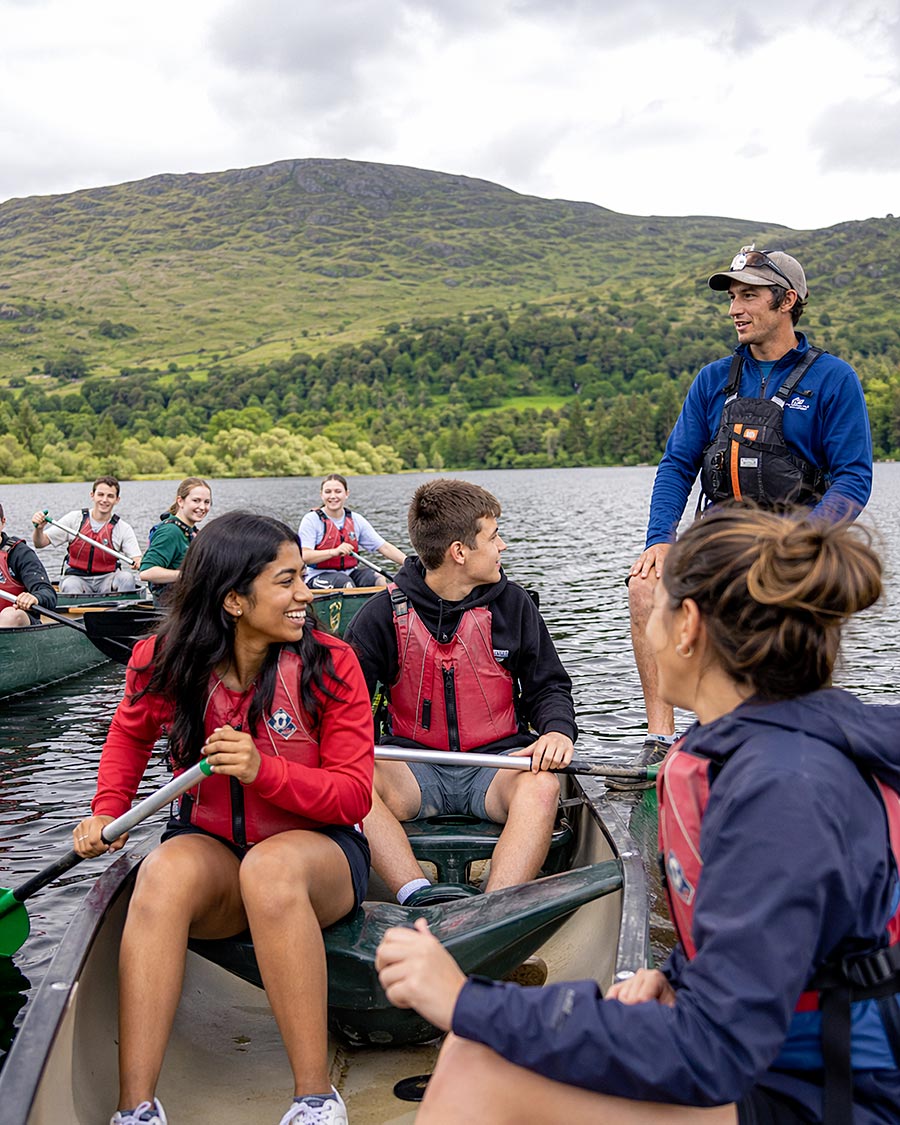 Un gruppo di giovani che indossano giubbotti di salvataggio siedono in canoa al Campo Avventura su un lago, sorridendo e ascoltando un istruttore in piedi in un'altra canoa, con colline verdi e un cielo nuvoloso sullo sfondo.