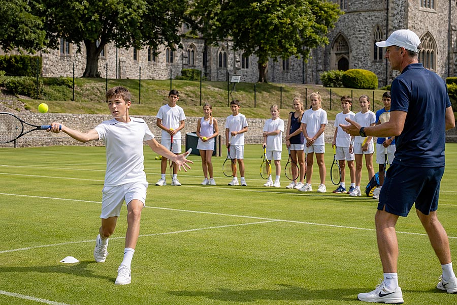 Un ragazzo vestito di bianco gioca a tennis su un campo in erba dell'Accademia del Tennis, brandendo la racchetta contro la palla di un allenatore adulto. Diversi bambini in bianco guardano con la racchetta in mano, mentre edifici in pietra e alberi fanno da sfondo.