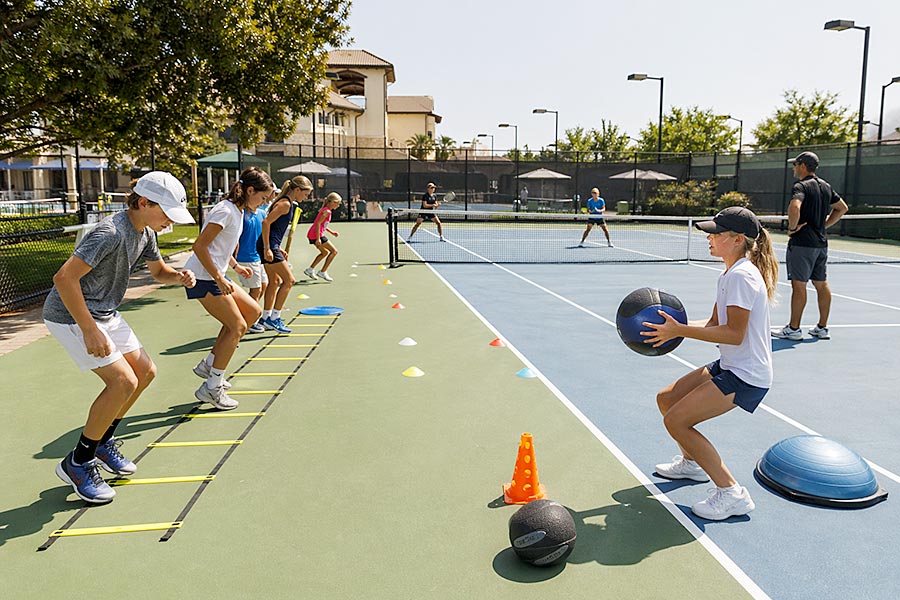 I bambini partecipano a un'esercitazione di fitness all'aperto presso la Tennis Academy, utilizzando scale di agilità, coni, palle mediche e scarpe da ginnastica per l'equilibrio su un campo da tennis, con gli allenatori che supervisionano e altri che giocano a tennis sullo sfondo.