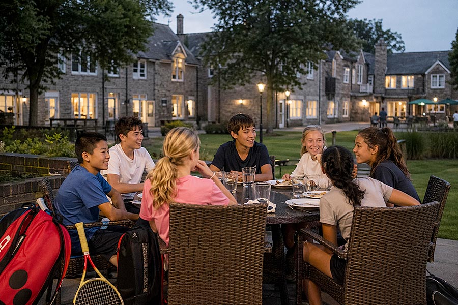 Un gruppo di sette adolescenti siede intorno a un tavolo all'aperto al tramonto, sorridendo e parlando mentre condividono il pasto davanti a un elegante edificio dell'Accademia del Tennis. Le racchette da tennis sono appoggiate su alcune sedie.