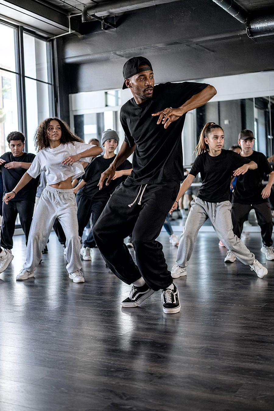 Un gruppo di persone in abiti casual, guidate da un istruttore, si esercita in un esercizio di danza in una scuola di danza con grandi specchi e finestre.