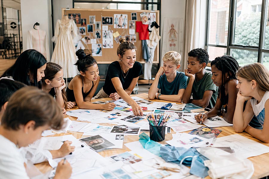 Un gruppo di studenti e un insegnante siedono attorno a un tavolo coperto di fogli, schizzi e materiale artistico, impegnati in un'attività creativa collaborativa in una luminosa e moderna aula della Dance Academy con disegni di moda esposti sullo sfondo.