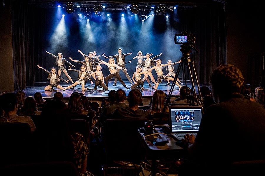 Un gruppo di ballerini dell'Accademia di danza si esibisce in un numero sincronizzato su un palcoscenico con luci scenografiche, mentre il pubblico osserva e una persona filma e monitora la performance da una postazione di controllo in primo piano.