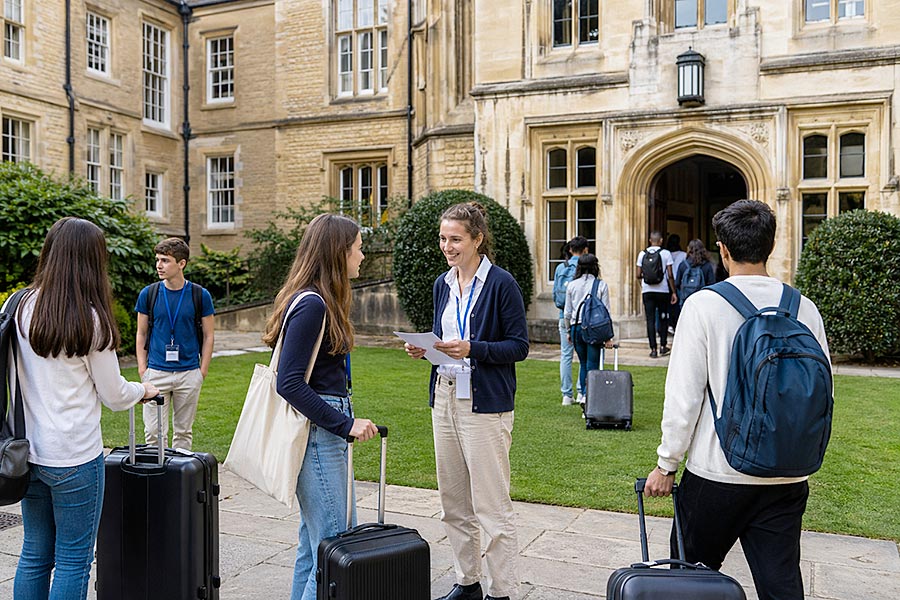 Un gruppo di studenti con le valigie sta in piedi e parla all'esterno di un edificio universitario storico, unendosi ad altri che stanno attraversando un ingresso ad arco - catturando l'eccitazione dell'arrivo per un Teen Program o un orientamento universitario.