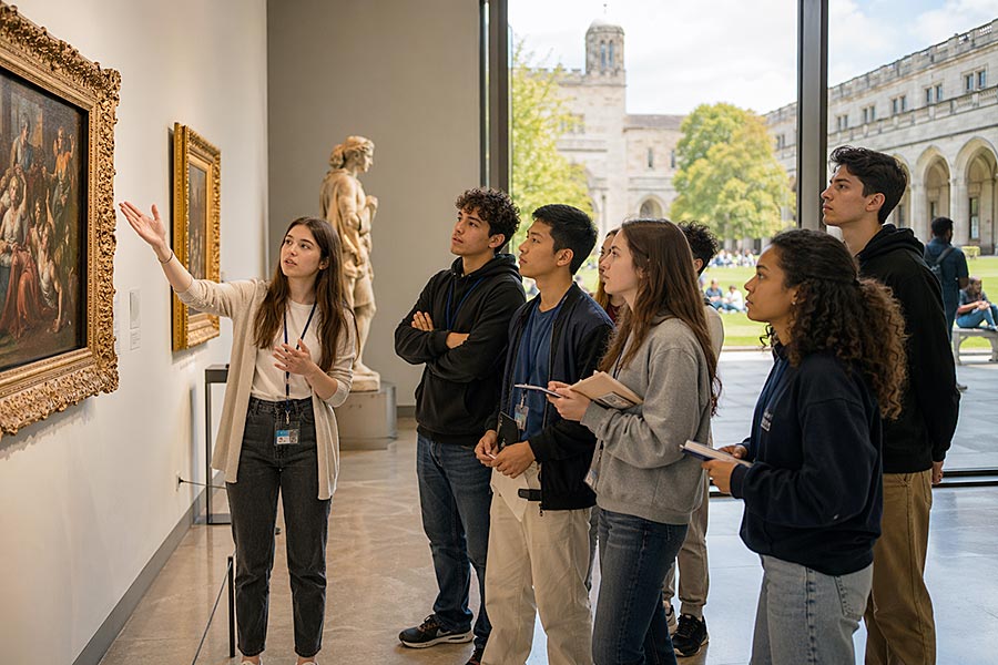 Una guida del museo fa un gesto verso un dipinto, spiegandolo a un gruppo di giovani adulti attenti che partecipano a un programma per adolescenti. Sono visibili opere d'arte classica e una statua, mentre all'esterno grandi finestre mostrano un campus universitario.