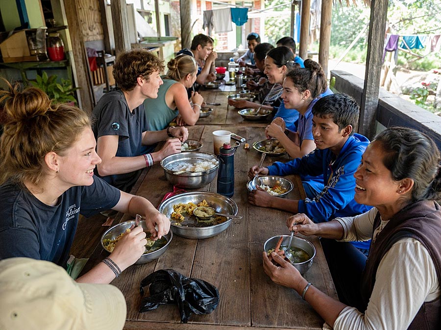 Un gruppo di persone siede a un lungo tavolo di legno all'aperto, condividendo un pasto e sorridendo mentre conversano di programmi di volontariato. Ciotole e piatti di cibo sono sparsi sul tavolo, creando un'atmosfera vivace e amichevole.