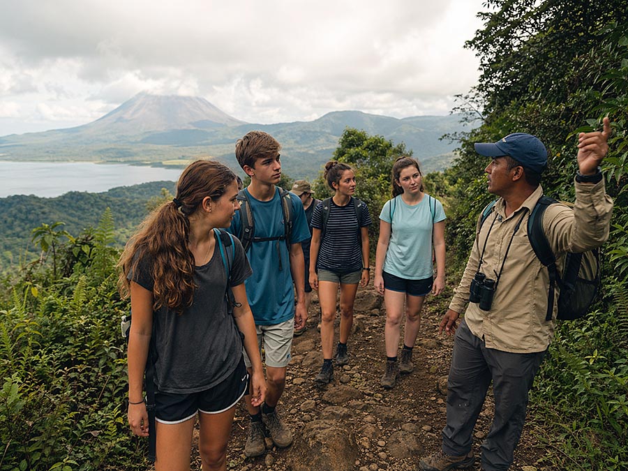 Un gruppo di quattro giovani adulti percorre un sentiero sterrato in una zona collinare e rigogliosa nell'ambito di progetti di volontariato, guidati da un istruttore che parla a gesti. Un vulcano e un lago sono visibili sullo sfondo sotto un cielo nuvoloso.