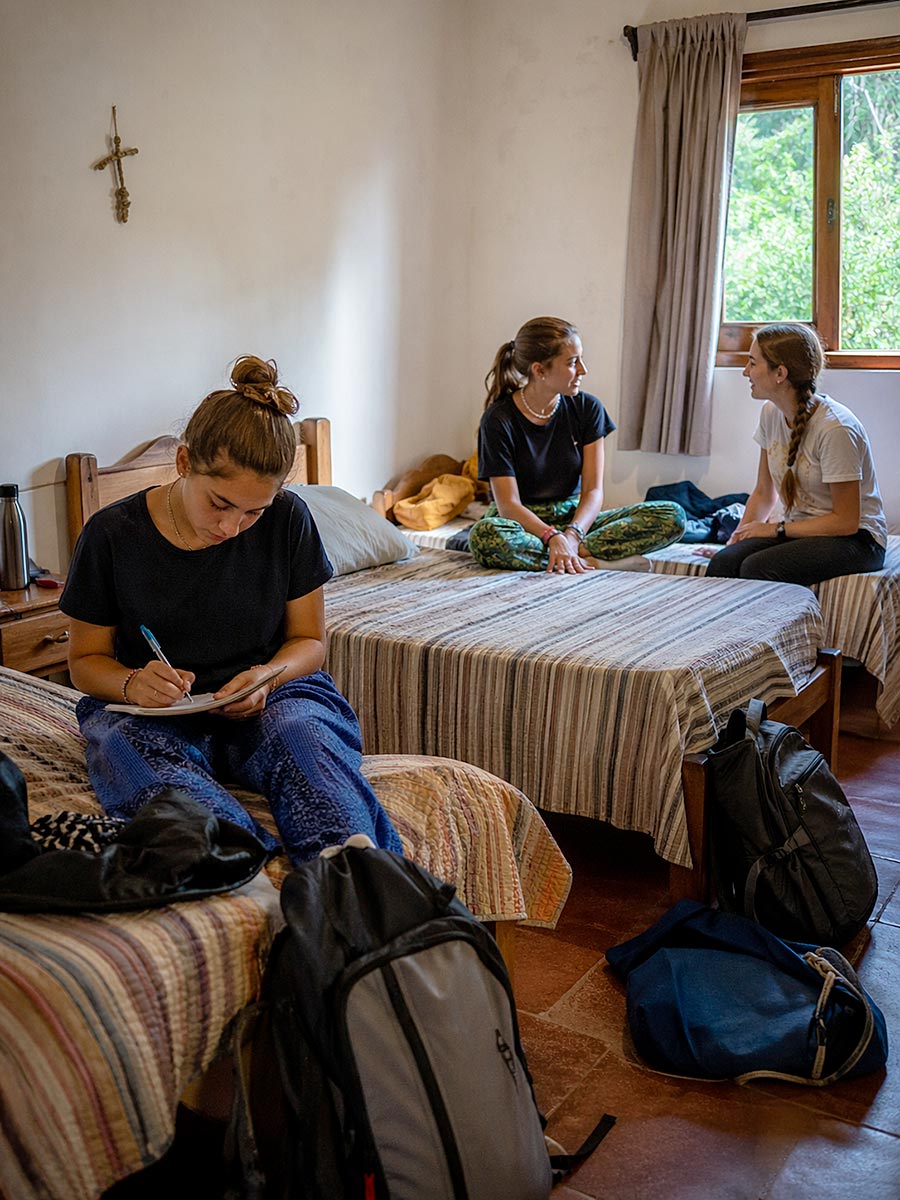 Tre giovani donne in abiti casual si rilassano in una camera da letto illuminata dal sole con copriletto a righe, chiacchierando e scrivendo. Gli zaini appoggiati sui letti indicano il loro coinvolgimento in programmi di volontariato, condividendo le storie del loro percorso di volontariato.