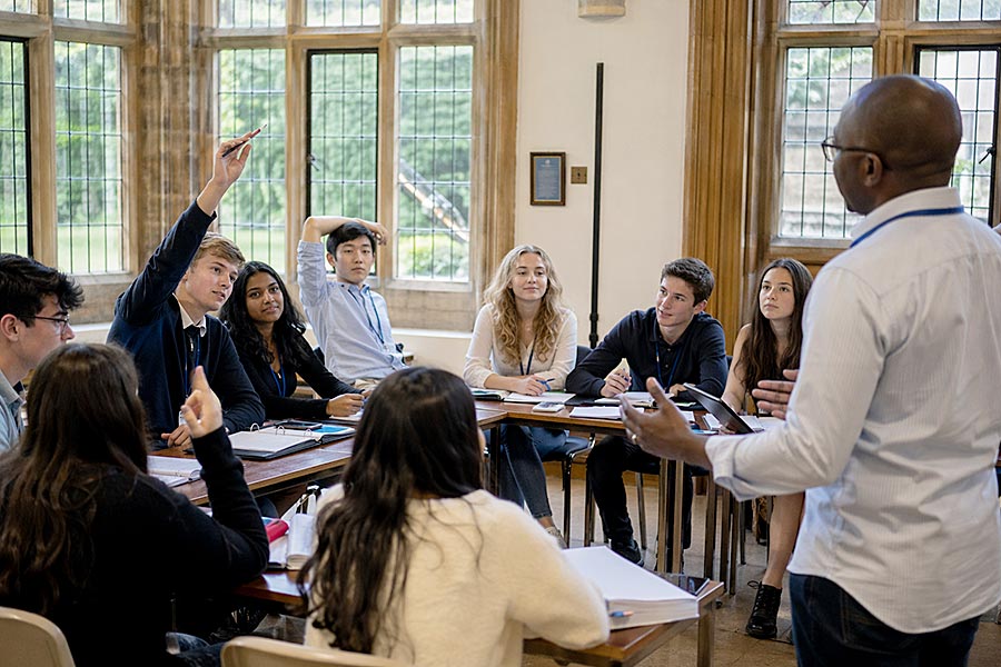 Un gruppo di studenti siede intorno a un tavolo, ascoltando attentamente un insegnante in piedi di fronte a loro. Uno studente ha la mano alzata, desideroso di partecipare ai corsi di leadership offerti in questa luminosa aula con grandi finestre.