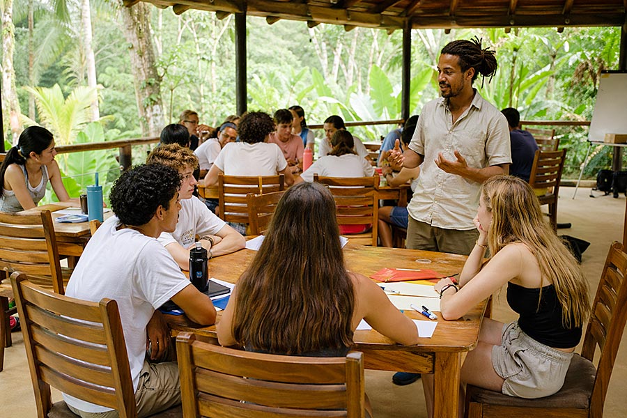 Un gruppo di studenti siede intorno a tavoli di legno all'aperto, circondati da una vegetazione lussureggiante, ascoltando un uomo che parla e gesticola durante un corso di leadership. Alcuni studenti scrivono mentre altri guardano e ascoltano con attenzione.