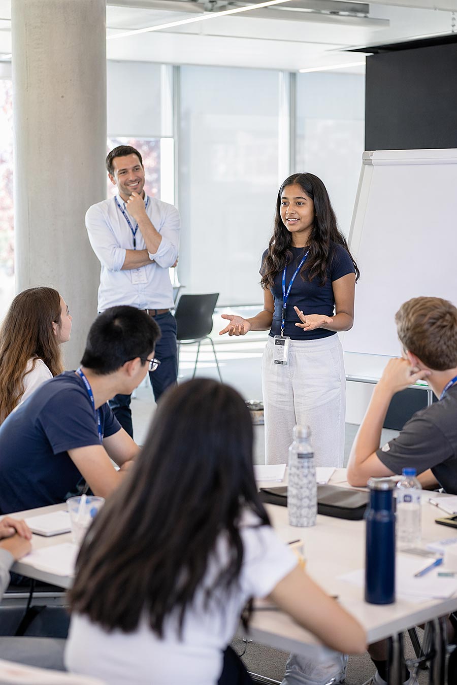 Una giovane donna è in piedi e parla a un gruppo di studenti seduti in un'aula luminosa durante Programmi di Leadership, mentre un uomo sta vicino sorridendo. Sui tavoli sono visibili una lavagna a fogli mobili e bottiglie d'acqua.