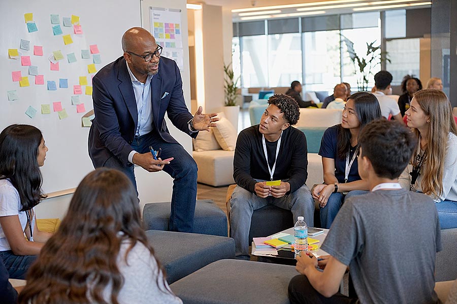 Un uomo in giacca e cravatta gesticola mentre parla a un gruppo eterogeneo di giovani adulti seduti in cerchio, impegnati in Programmi di Leadership. Note adesive colorate decorano la parete alle loro spalle in un moderno ufficio aperto.