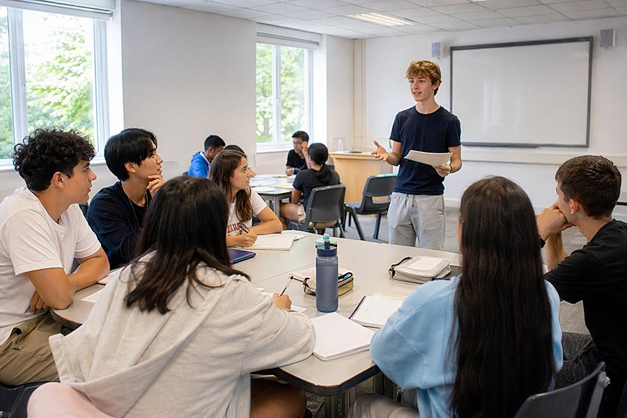 Un gruppo di studenti siede intorno a un tavolo in un'aula, ascoltando uno studente in piedi che parla davanti a sé durante una preparazione alla certificazione linguistica. Sul tavolo ci sono quaderni e bottiglie d'acqua; sullo sfondo una lavagna e delle finestre.