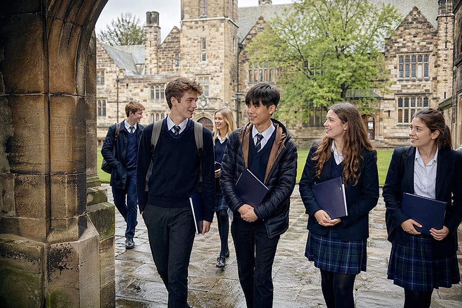 Un gruppo di studenti liceali in uniforme cammina e parla insieme all'esterno di uno storico edificio scolastico in pietra in una giornata di pioggia, portando cartelle e zaini durante il programma di lingua italiana.