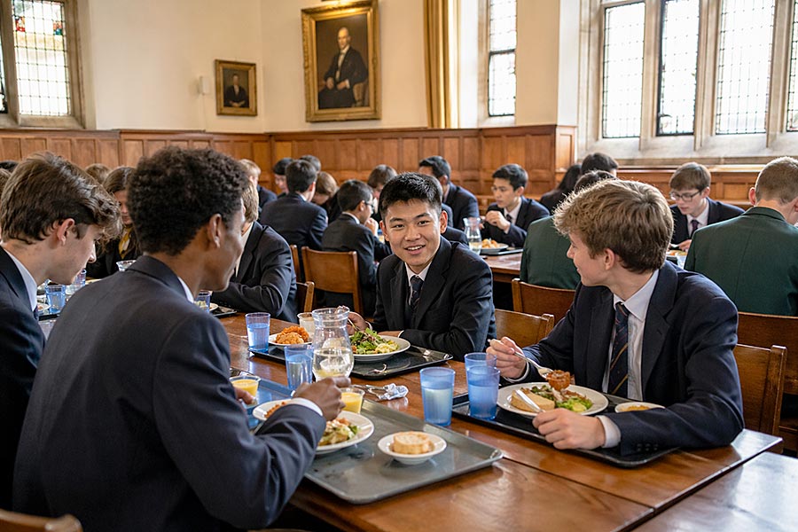 Studenti in uniforme scolastica siedono a lunghi tavoli di legno in una sala da pranzo durante il semestre, pranzando e parlando. La sala ha grandi finestre, pareti rivestite di legno e un ritratto appeso alla parete.