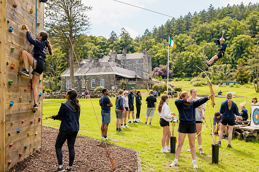 Un gruppo di adolescenti si diverte con le attività all'aperto del Campo Avventura, tra cui l'arrampicata, il tiro con l'arco e la teleferica, in un campo erboso vicino a un grande edificio in pietra circondato da alberi.